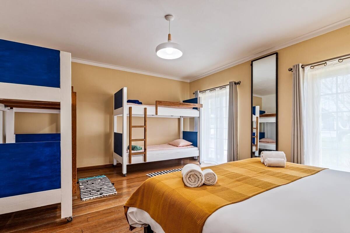 Una habitación con literas, una cama con una manta amarilla, una escalera de madera, una alfombra pequeña y un espejo grande.