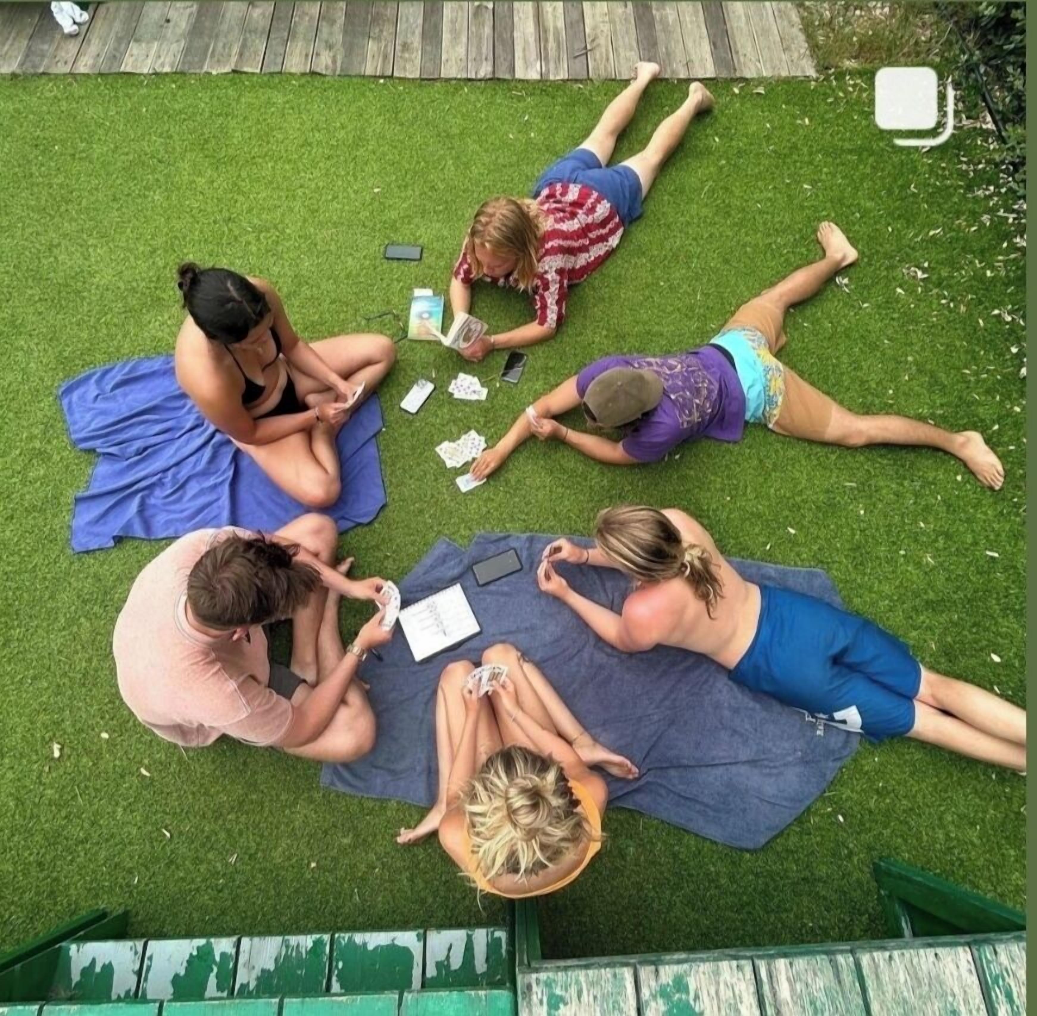 A group of people lying on a blue mat on artificial grass, engaged in various activities like reading and playing cards.