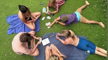 A group of people lying on a blue mat on artificial grass, engaged in various activities like reading and playing cards.