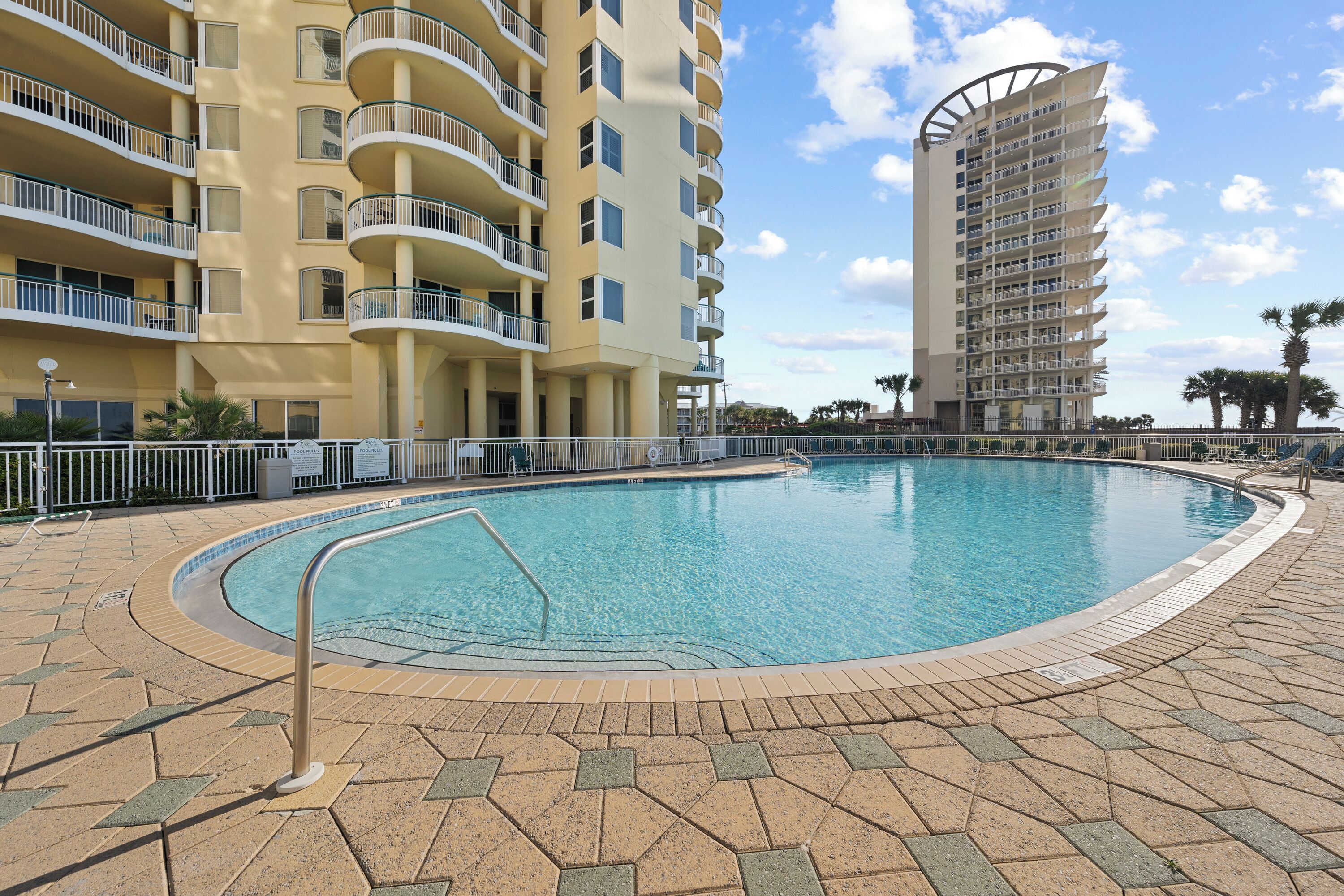 A swimming pool surrounded by a tiled deck, with a multi-story building featuring balconies in the background.