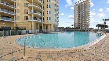 A swimming pool surrounded by a tiled deck, with a multi-story building featuring balconies in the background.