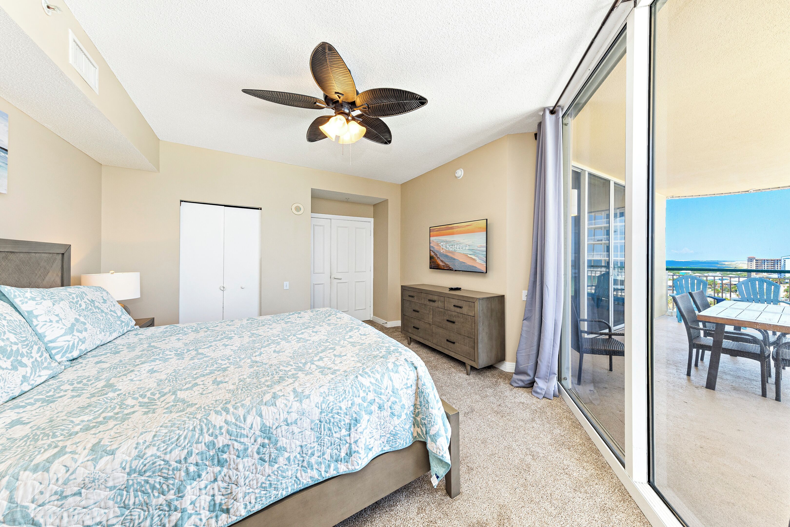 A bedroom with a bed, a ceiling fan, a dresser, and a balcony with outdoor furniture.