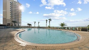 A swimming pool with a curved edge, surrounded by a tiled deck and palm trees. - Beach Colony E10B (Pensacola)