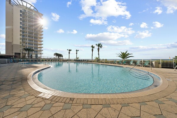 A swimming pool with a curved edge, surrounded by a tiled deck and palm trees. - Beach Colony E10B (Pensacola)