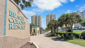 A pathway leading to a building labeled "Beach Colony Resort" with tall residential buildings in the background. - Beach Colony E10B (Pensacola)
