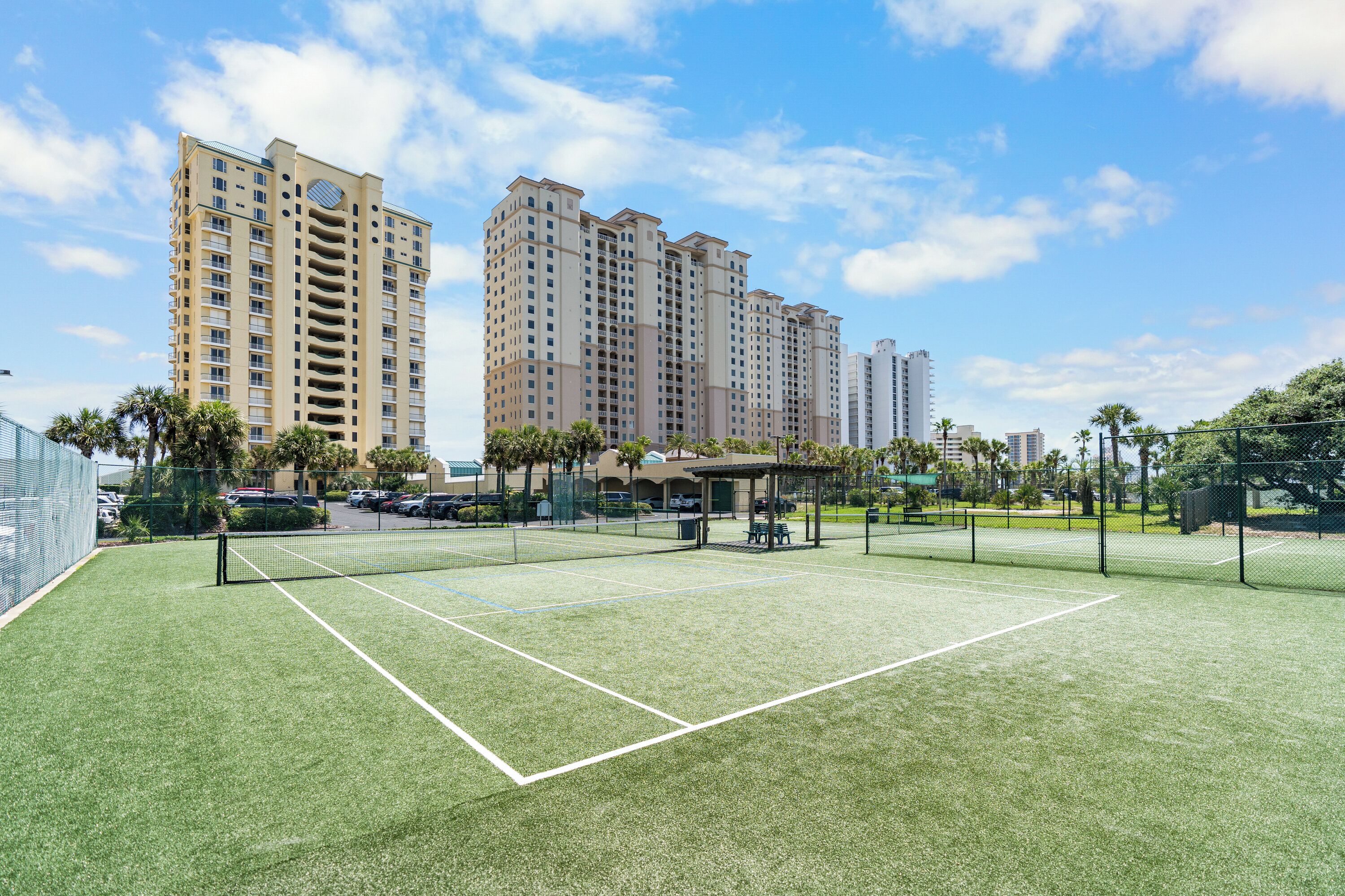 A tennis court with a net, surrounded by a fence, and tall residential buildings in the background.