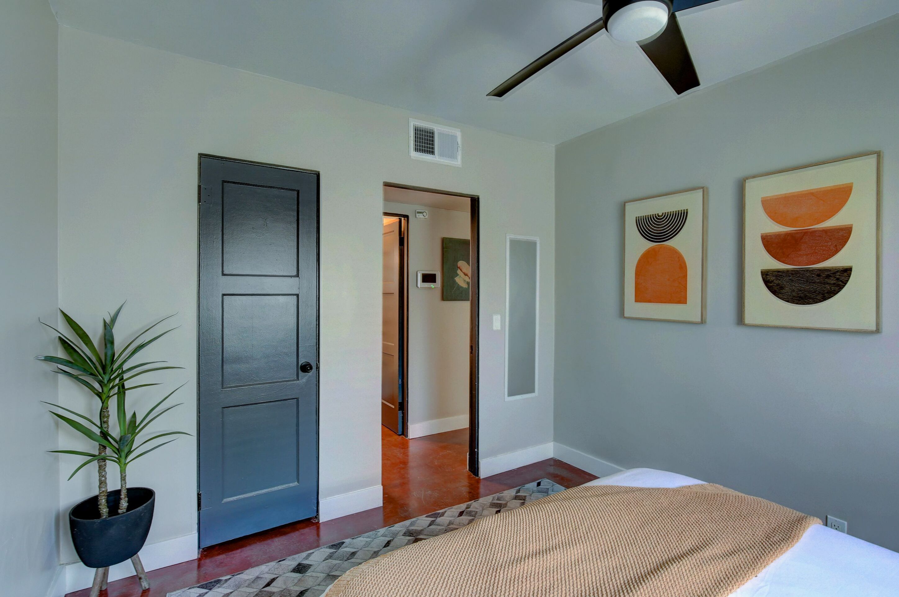 A bedroom with a bed, a potted plant, a ceiling fan, and artwork on the wall.