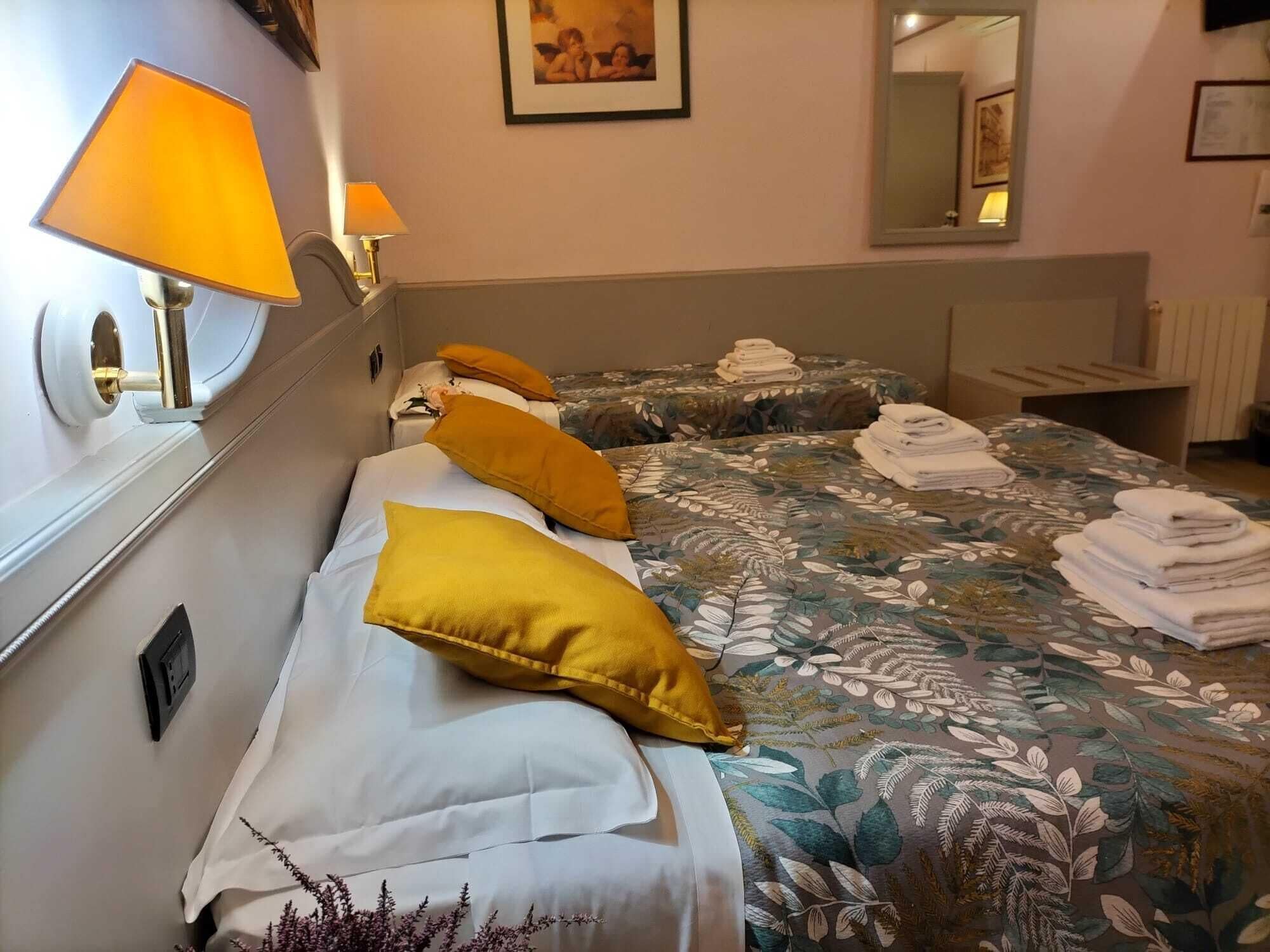 Une chambre d’hôtel avec deux lits, chacun recouvert d’une courtepointe à motifs floraux et agrémenté d’une serviette pliée sur le côté. Deux lampes murales aux abat-jour jaunes sont fixées au mur.