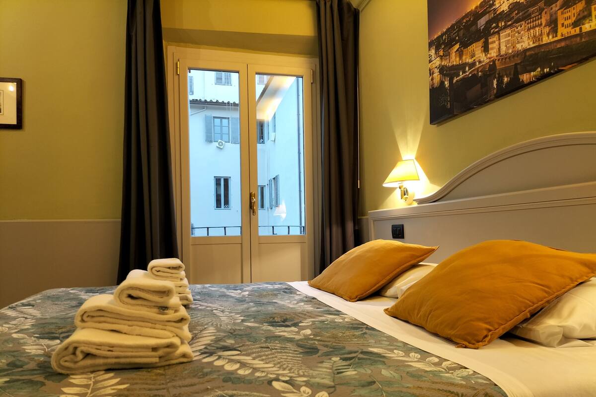 A hotel room with a bed, a folded towel, a lamp, and a view of buildings through a door.