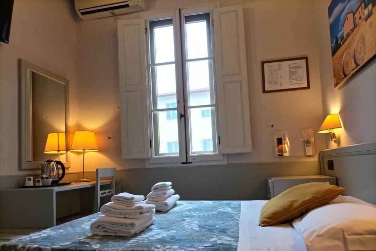 A hotel room with a bed, a desk with a lamp, a chair, a window with shutters, and a wall-mounted air conditioner.