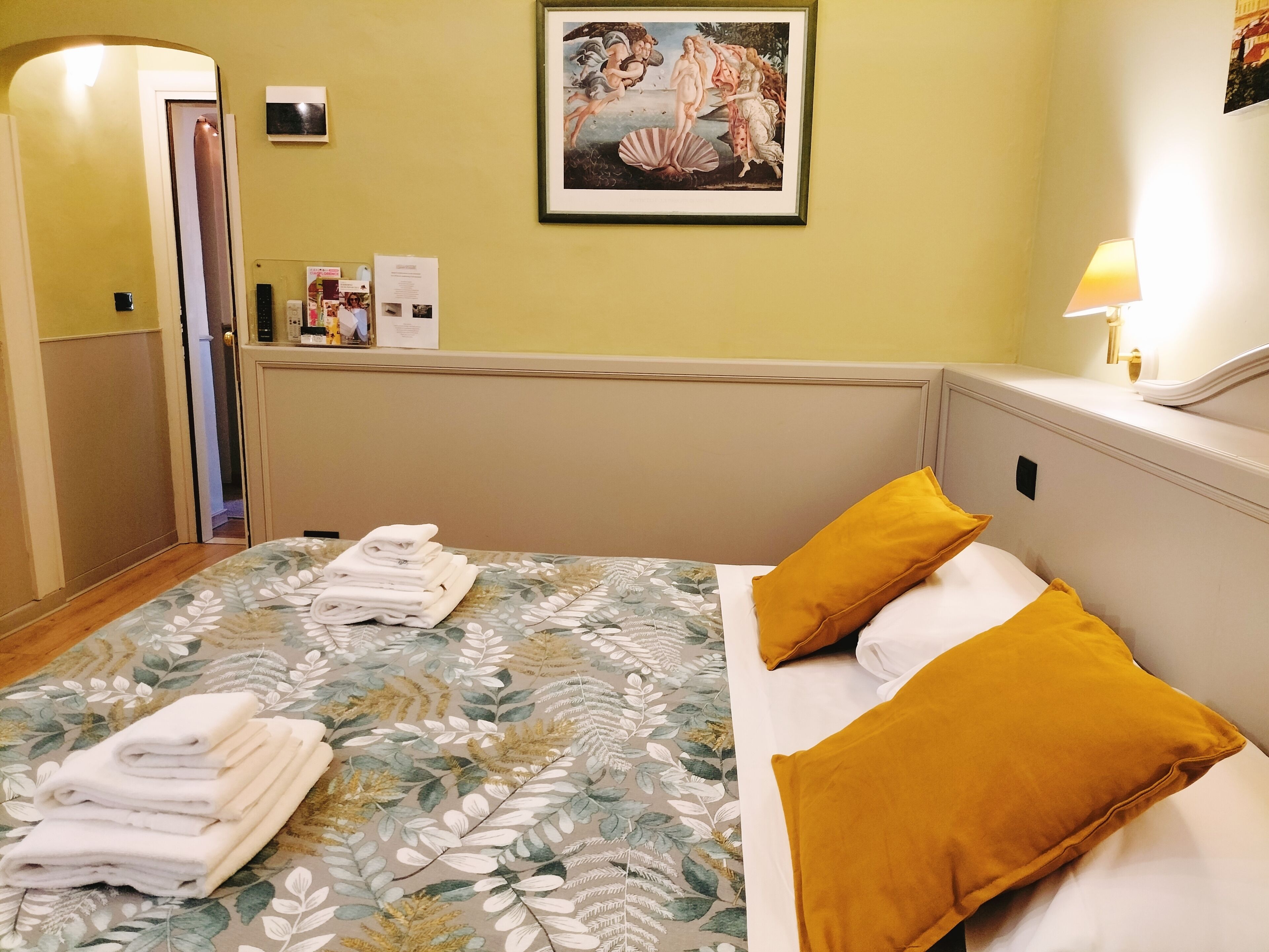 A hotel room with a bed, a door, a framed picture on the wall, and a lamp on the nightstand.