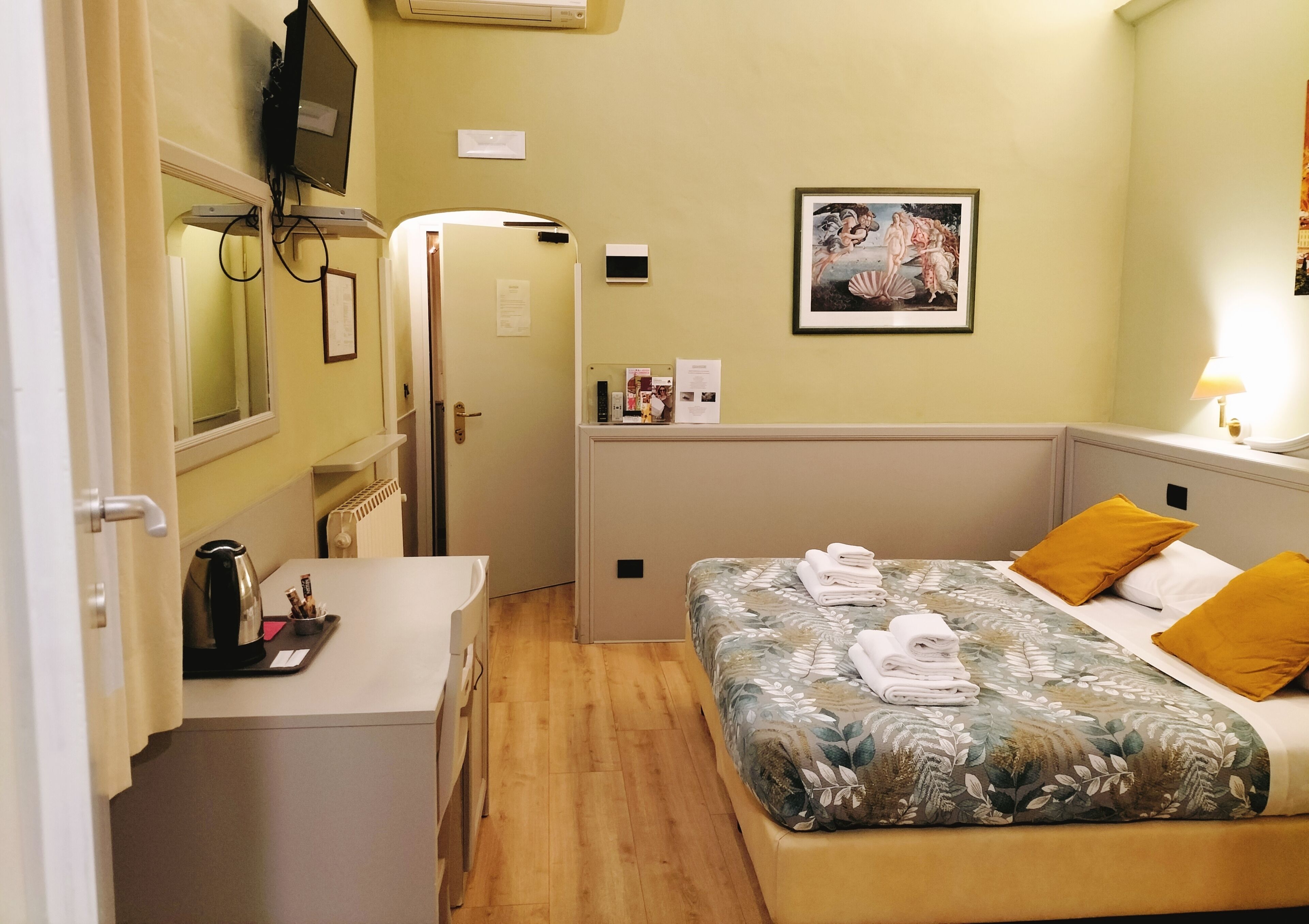 Habitación de hotel con cama, escritorio, silla, radiador, televisor y un cuadro en la pared.