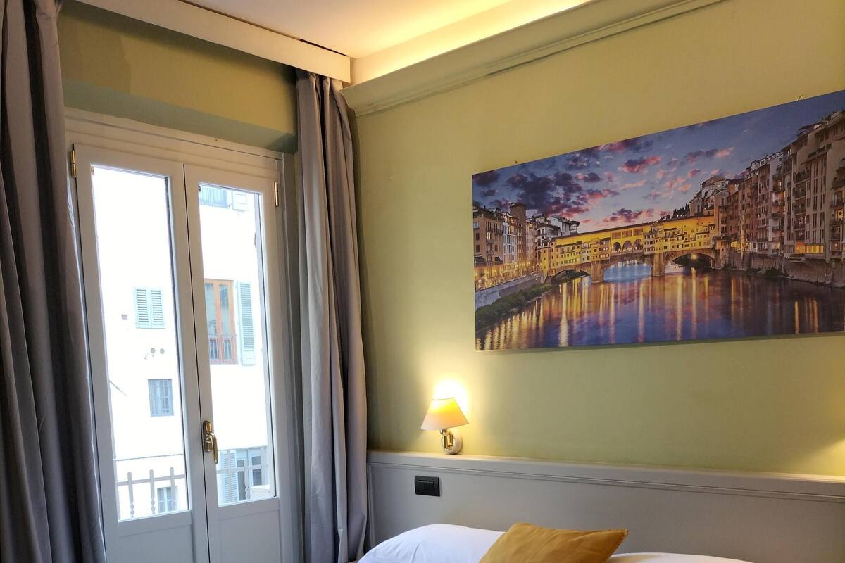 A hotel room with a bed, a painting of a cityscape, a lamp, and a window with curtains.