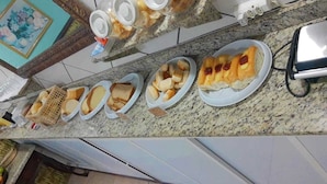 A kitchen counter with various food items and containers, including plates with bread and other baked goods, a basket, and a toaster. - POUSADA PONTA DO ARPAO (Mangaratiba)
