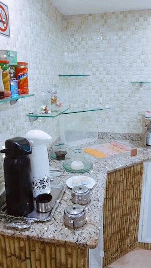 Una cocina con encimera de granito, cafetera, hervidor y repisa de vidrio con diversos objetos.