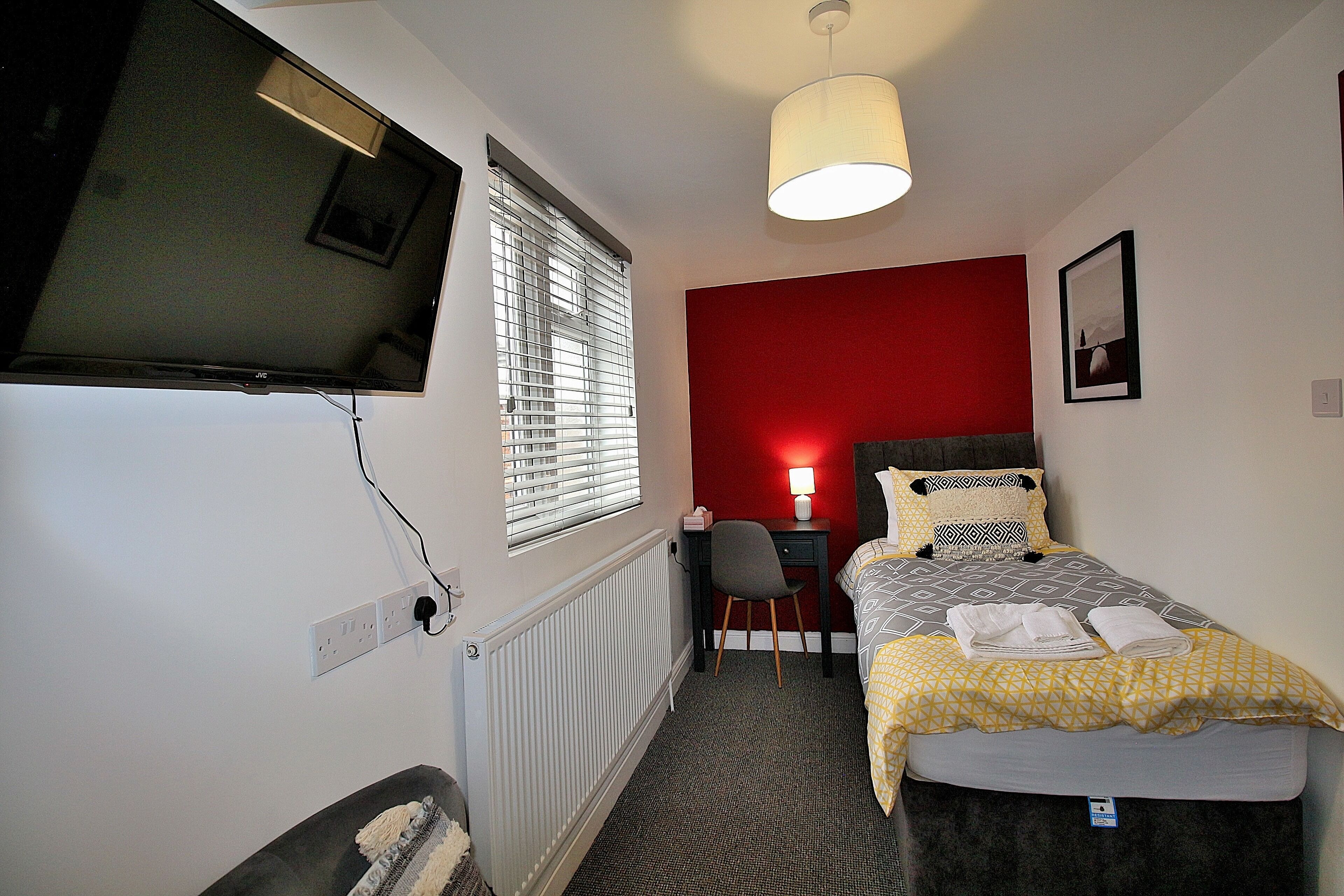 A small, compact room with a bed, a desk with a chair, a TV mounted on the wall, and a window with blinds.