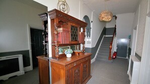 Une bibliothèque en bois remplie de livres et d’objets décoratifs, un escalier surmonté d’un lustre, et un extincteur.