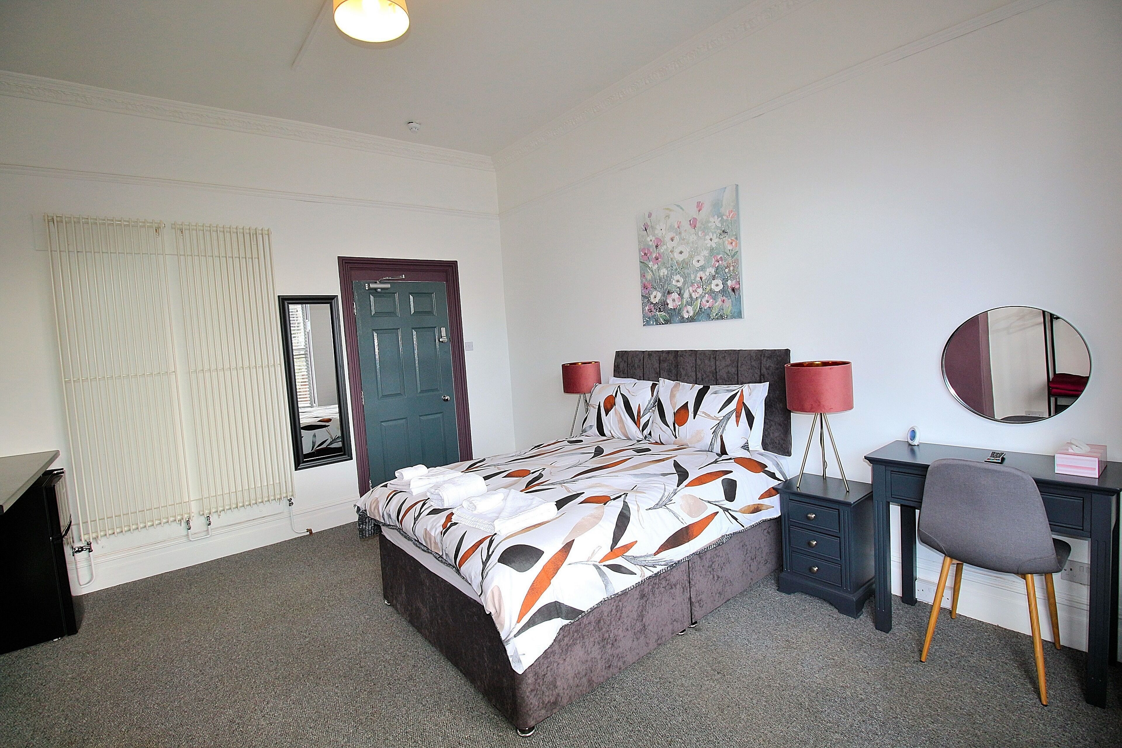 A bedroom with a bed, a desk, a chair, a mirror, a radiator, and a door.