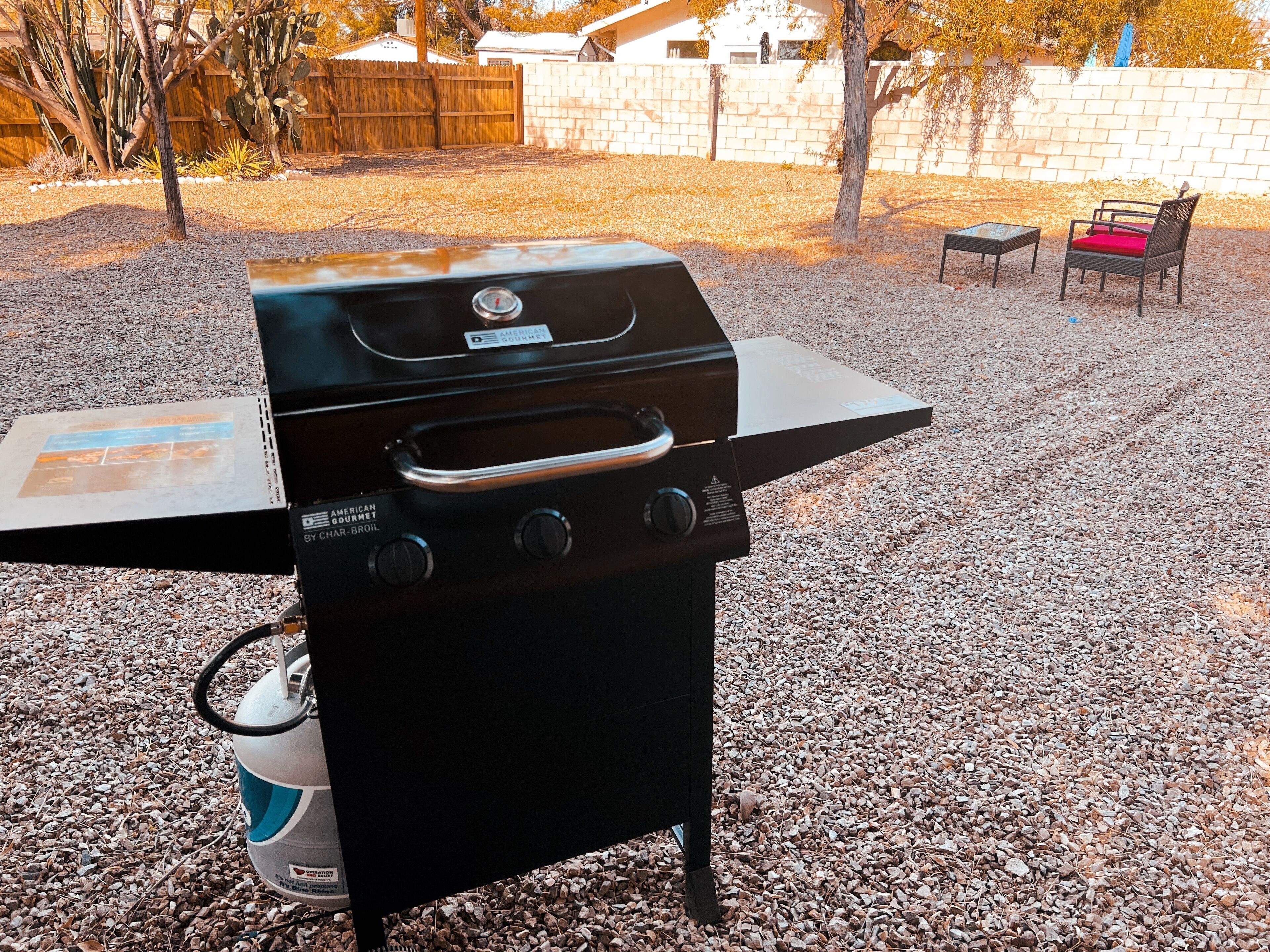 A black outdoor grill with a gas tank and a control panel, set on a gravel surface with a wooden fence and a brick wall in the background.