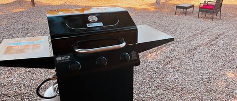 A black outdoor grill with a gas tank and a control panel, set on a gravel surface with a wooden fence and a brick wall in the background.