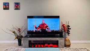 Een flatscreen televisie op een standaard in een woonkamer met een kamerbreed tapijt, een vaas met bloemen en twee ingelijste foto's aan de muur.