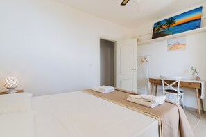 Ett hotellrum med en säng, ett skrivbord, en stol, en lampa och en tavla som föreställer en strandvy.