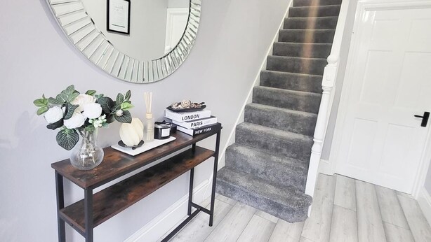 Una escalera con alfombra gris, una puerta blanca y una consola con un espejo, libros y un florero con flores.