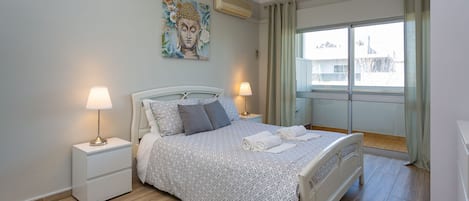 A bedroom with a bed, two bedside tables, a lamp, a painting, and a window with curtains.