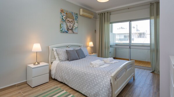 A bedroom with a bed, two bedside tables, a lamp, a painting, and a window with curtains.