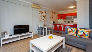 A modern living room with a TV, white furniture, a dining area, and a kitchen with red cabinets.