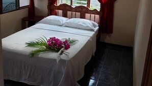 A single bed with a white bedspread and pillows, a wooden headboard, red curtains, and an air conditioning unit above the window. - Cristina's (Cóbano)