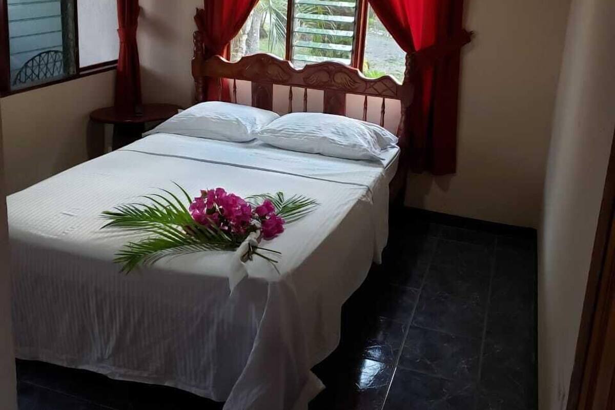 A single bed with a white bedspread and pillows, a wooden headboard, red curtains, and an air conditioning unit above the window.