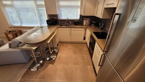 A modern kitchen with a built-in island, stainless steel appliances, and a seating area with a sofa and bar stools. - The Mumbles - Bungalow - 2 Bedrooms - Pet Friendly (Swansea)