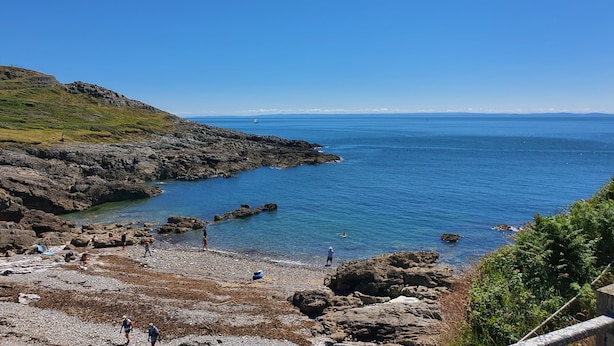 A rocky coastal area with clear blue water, a few people walking, and a distant sailboat. - The Mumbles - Bungalow - 2 Bedrooms - Pet Friendly (Swansea)