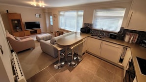 A modern kitchen with a central island, built-in appliances, and a dining area with a sofa and ottoman. - The Mumbles - Bungalow - 2 Bedrooms - Pet Friendly (Swansea)