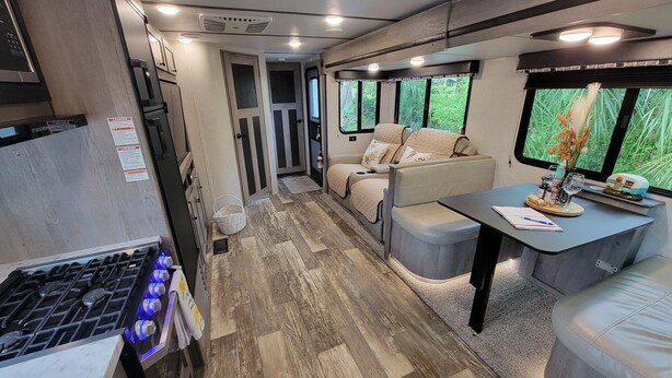 A compact RV interior with a kitchenette, seating area, and large windows offering a view of greenery. - New little getaway (Fernandina Beach)