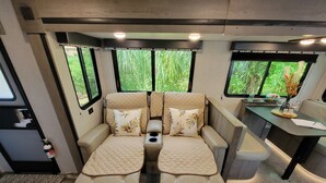 A cozy RV interior with two cushioned seating areas, a small table with a tray, and large windows offering a view of greenery. - New little getaway (Fernandina Beach)