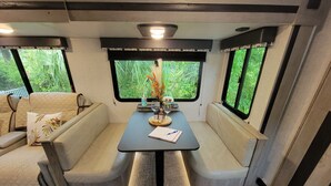 A cozy RV interior with a dining area, cushioned seating, and large windows offering a view of greenery. - New little getaway (Fernandina Beach)