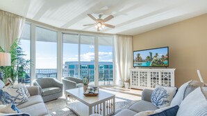 A spacious living room with a large window, a ceiling fan, and a flat-screen TV. - Two Bedroom Penthouse with Balcony Overlooking Pool Decks and Gulf. Complimentary Boat Slip! (Orange Beach)