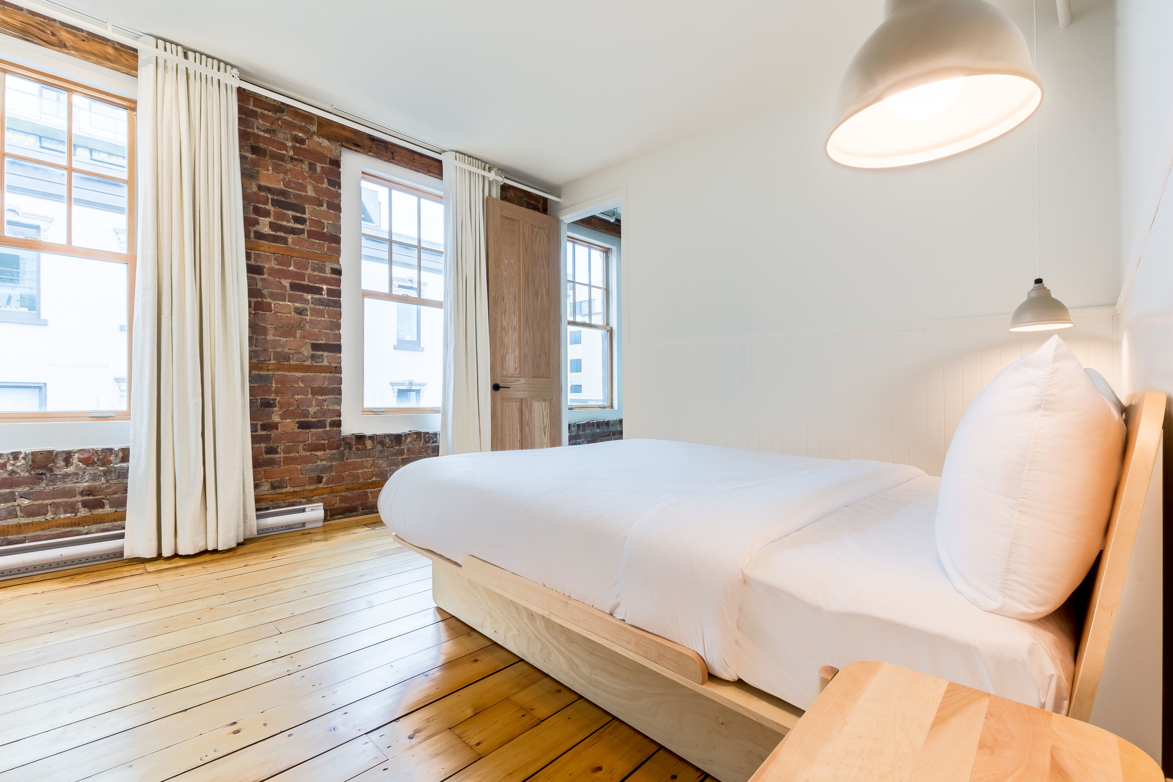A bedroom with a wooden floor, a bed with white bedding, a brick wall, and large windows with curtains.