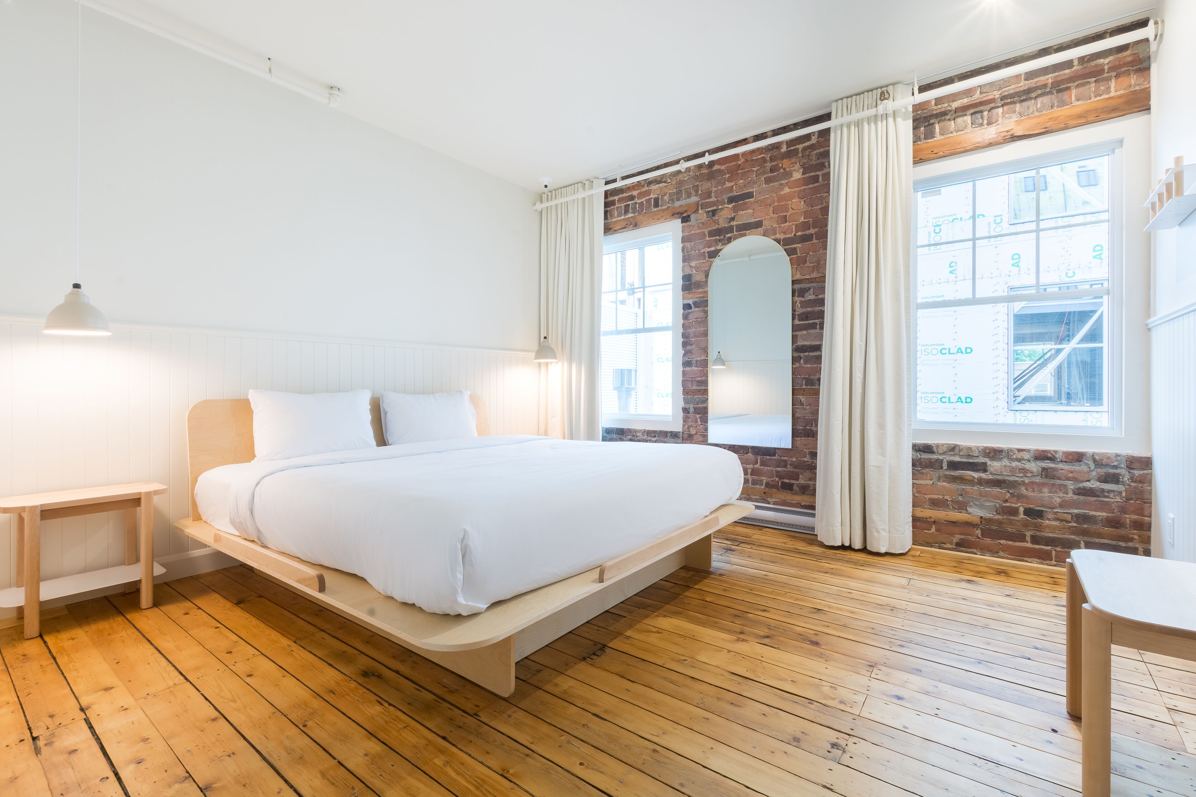 Un dormitorio con suelo de madera, una cama con ropa de cama blanca, una mesita de noche, un espejo y una ventana con cortinas.