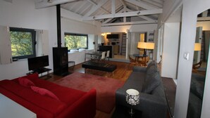 A spacious living area with a red sofa, a grey sofa, and a black ottoman. There is a television and a fireplace. The room has large windows and a high ceiling with exposed beams. - Casa do Rio Vez - LOFT IN RIVERFRONT (Arcos de Valdevez)