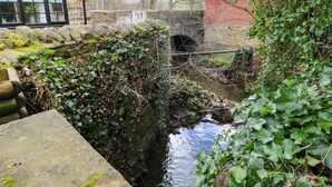 Um canal de água estreito com uma ponte de pedra, rodeado por vegetação e um edifício de tijolos.