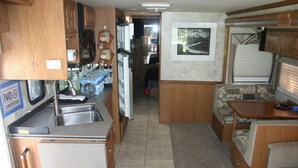Een compacte camperkeuken met een spoelbak, fornuis en koelkast. Er is een eethoek met een tafel en een bank.