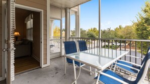 A covered patio with a glass table and blue chairs, a door leading to a room with a lamp, and a view of a street and trees. - 280-15 Woodlands Way (Calabash)