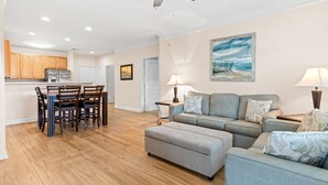 A spacious living room with a grey sofa, a dining area with wooden chairs, and a kitchen with wooden cabinets. - 280-15 Woodlands Way (Calabash)