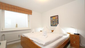 Ein ordentlich bezogenes Bett mit weißen Leinen und Handtüchern, ein Nachttisch aus Holz mit einer Lampe, eine Heizung und ein Fenster mit Jalousien.