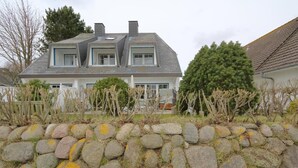 Ein zweistöckiges Haus mit einer Steinmauer im Vordergrund, einem umzäunten Garten und einem großen Nadelbaum.