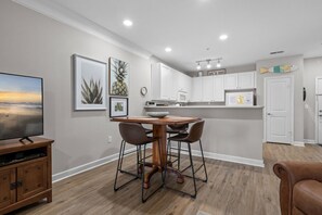A modern kitchen with white cabinets, a dining table with chairs, and a TV on the left. - Crow Creek Condo minutes from Sunset Beach (Calabash)