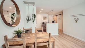Uma área de refeições moderna com uma mesa e cadeiras de madeira, um espelho grande, plantas penduradas e uma cozinha ao fundo.